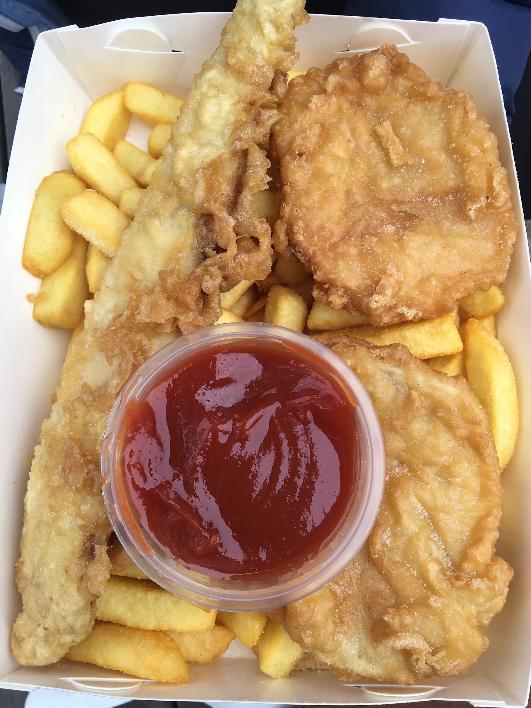 A box of battered fish, potato cakes and chips with a tub of tomato sauce